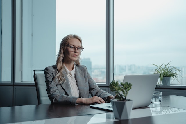A businesswoman in a grey office with a city behind her looking at a computer